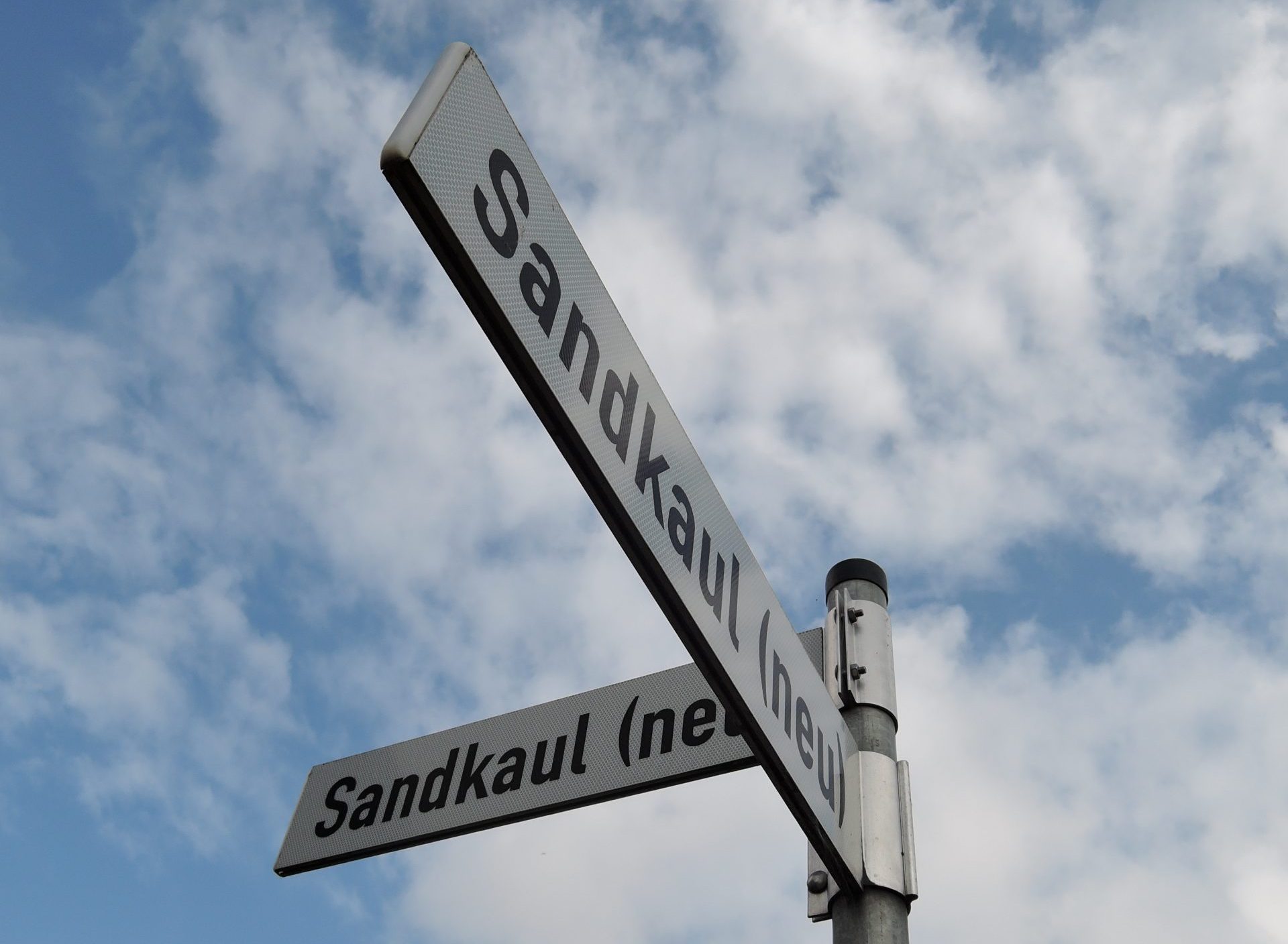 Straßenschild mit dem Namen "Sandkaul (neu)". Im Hintergrund ist ein blauer Himmel mit weißen Wolken zu sehen.