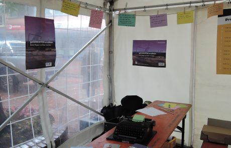 Blick in den Pavillon: Ein Tisch mit Schreibmaschine und Werbematerialien, Plakate und bunte Zettel mit Zitaten sind zu sehen.