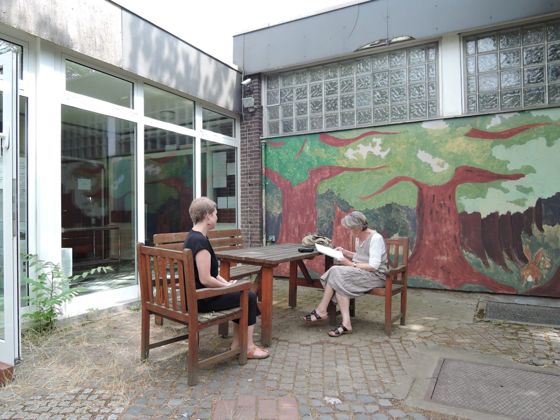 Ulrike Thiel und eine Mitarbeiterin des Projektteams "geSCHICHTEN Rheinisches Revier" sitzen an einem Tisch auf dem Schulhof in Alt-Morschenich. Frau Thiel liest etwas vor.